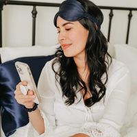 Woman looking at her phone in bed with a navy mulberry silk sleep mask pushed up on her head